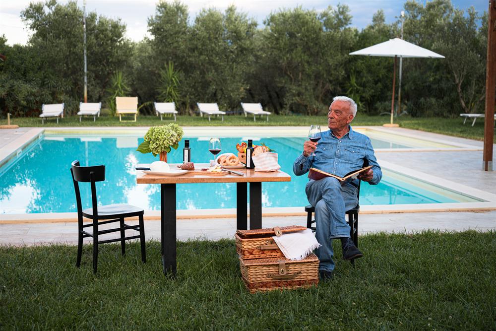 Un signore beve un calice di vino a un tavolo apparecchiato a bordo piscina, con olio, pane e salumi