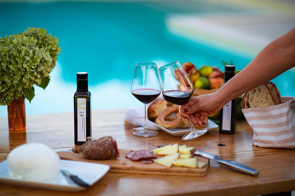 Bottiglie di olio extravergine, calici di vino rosso, salumi, formaggi e pane vicino alla piscina