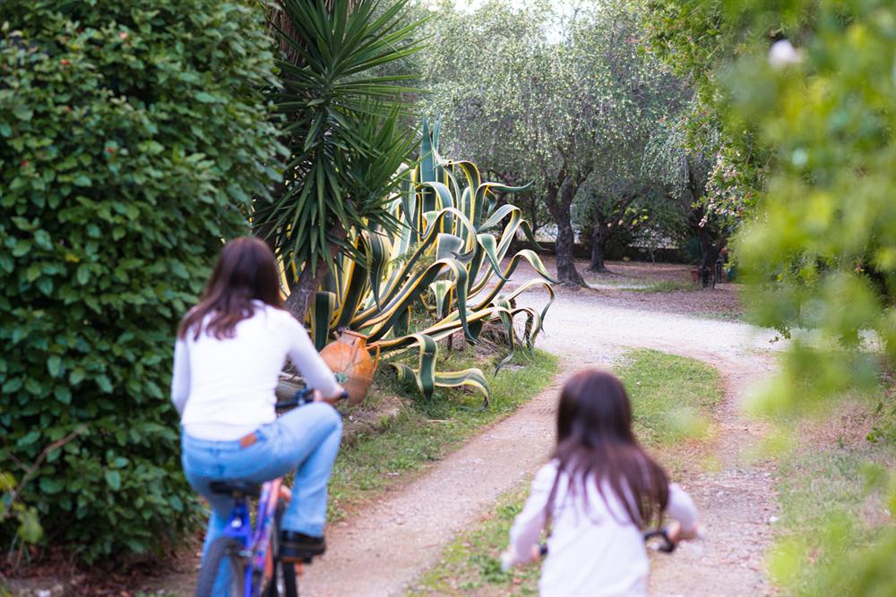 Due bambine in bicicletta lungo il viale di ghiaia tra agavi e ulivi
