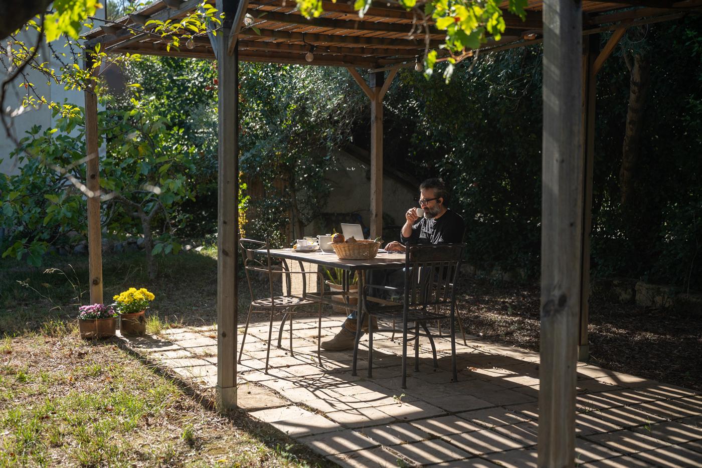 Un uomo fa colazione al tavolo sotto la pergola in giardino, tra gli ulivi